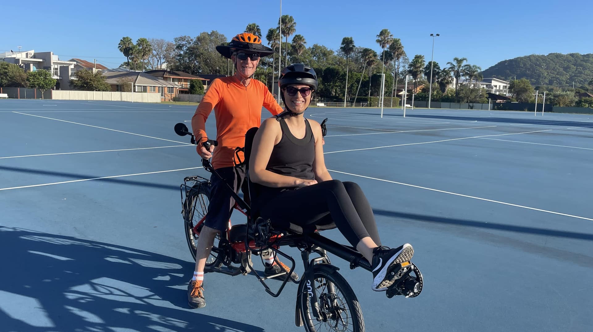 A pilot in orange stands behind a rider seated on the front of a tandem bicycle.