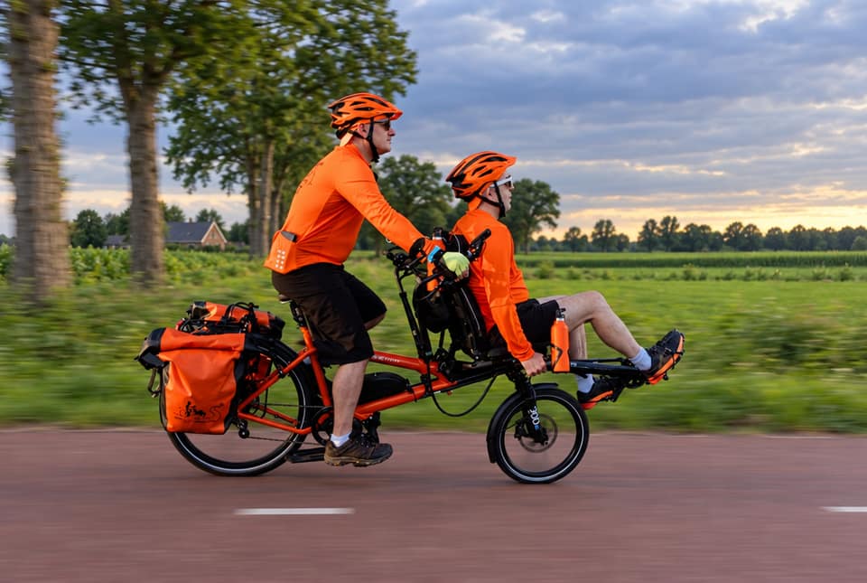 A pilot and a reclined front rider, both in orange, riding a loaded Hase Pino along a tree-lined country road.