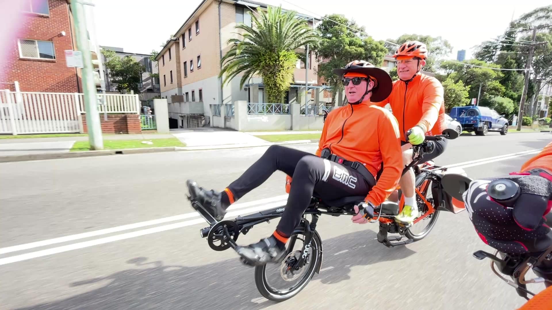 A pilot steering a Pino tandem along a suburban street, the rider reclined up front, both in orange.