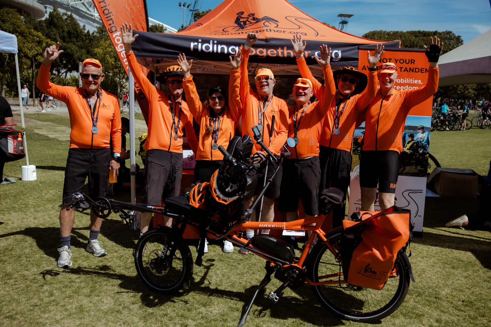 Seven Riding Together volunteers in orange jerseys with medals, arms raised at the team tent behind a Pino tandem.